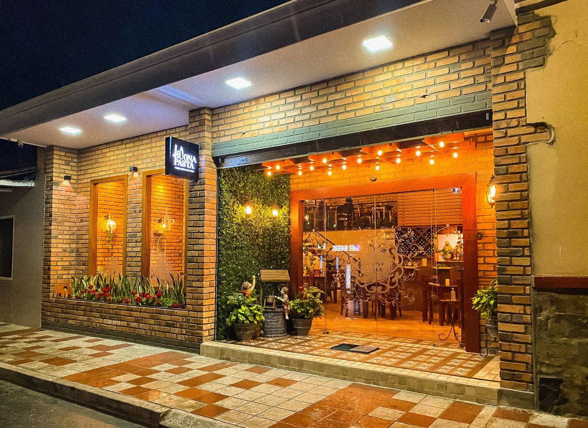 Fachada de La Buona Pasta con paredes de ladrillo, luces de hilo y entrada calida de madera